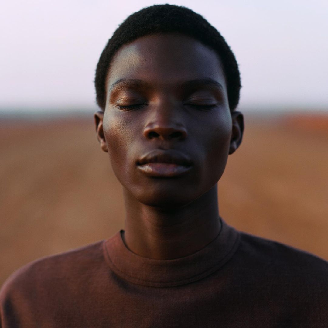 Close-up portrait of a young, dark-skinned adult with closed eyes, wearing a brown t-shirt, against a blurred horizon and tan background.