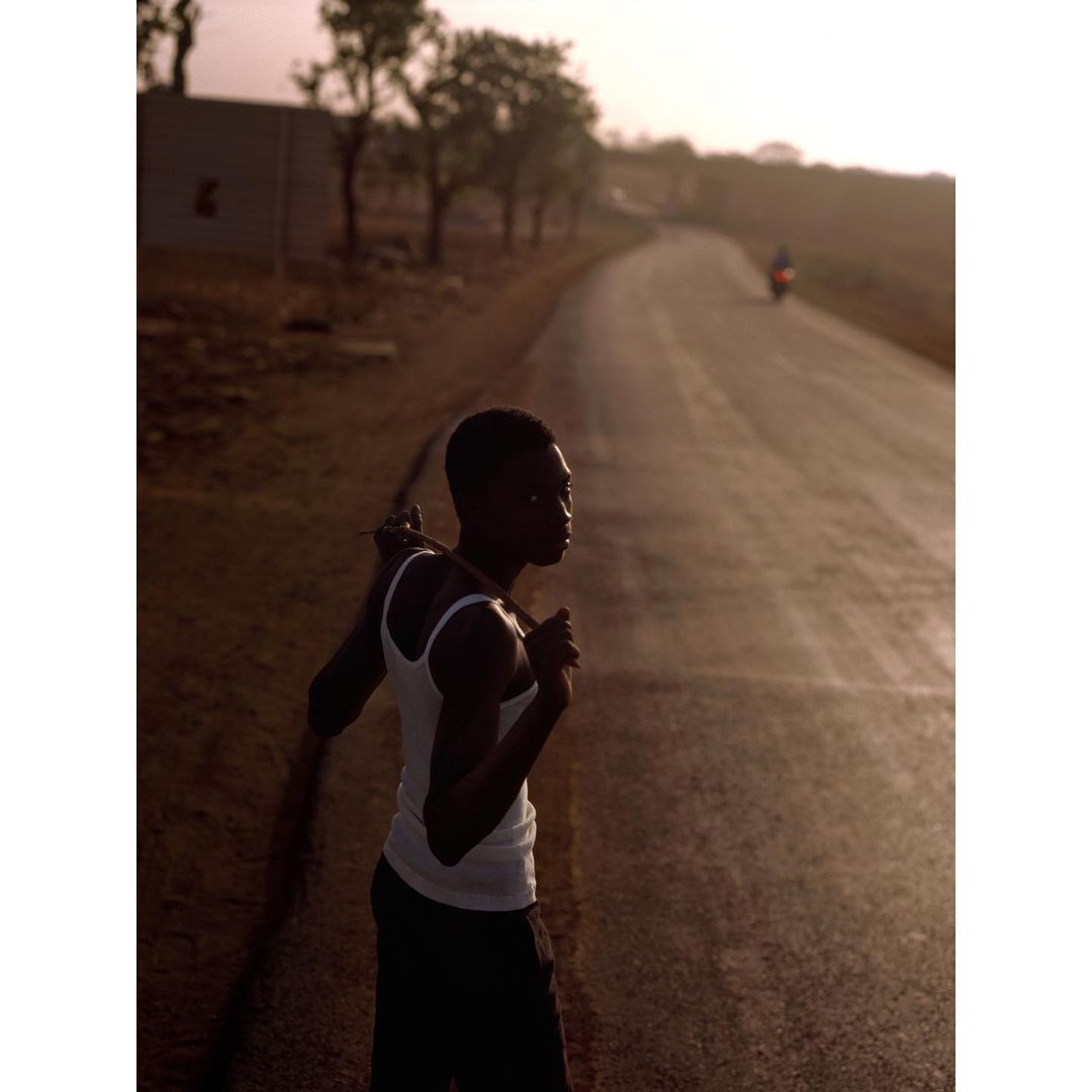 Young Black man in white tank top stands on a sunlit road near trees, creating a silhouetted effect, with a distant motorcyclist.