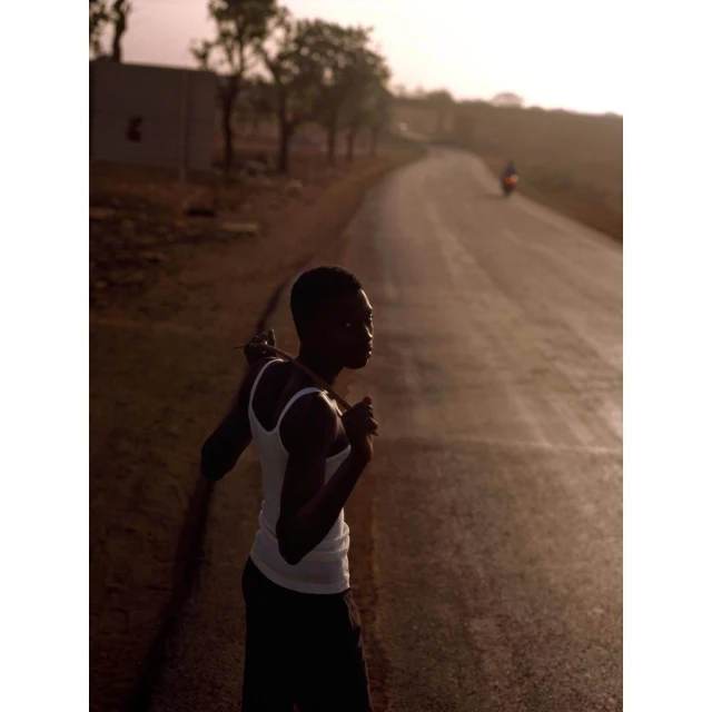 Young Black man in white tank top stands on a sunlit road near trees, creating a silhouetted effect, with a distant motorcyclist.