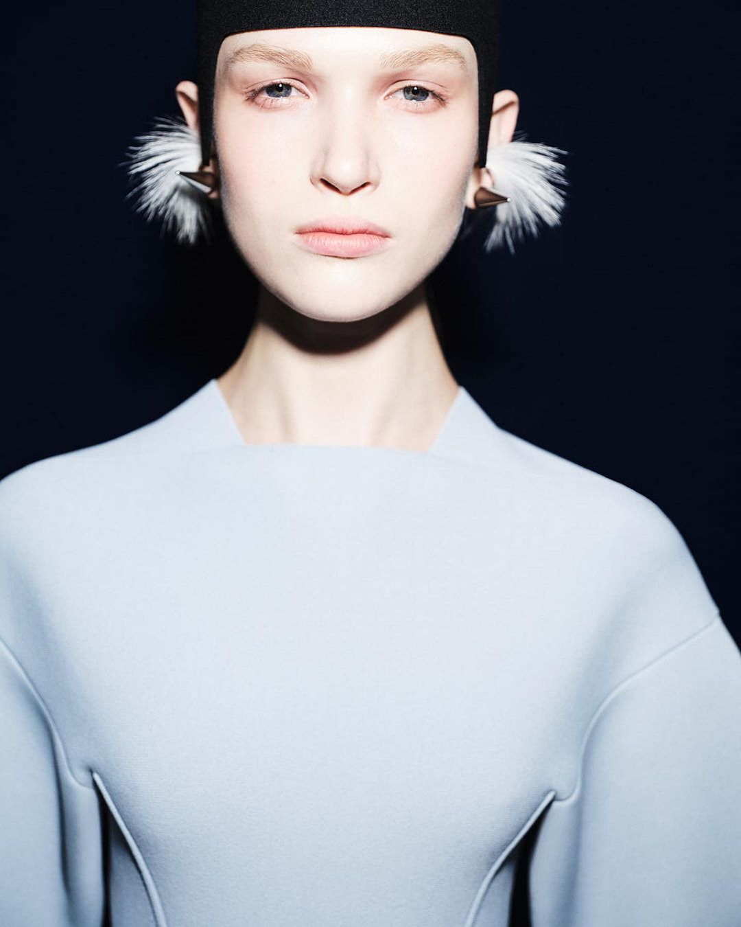 Close-up shot of a fair-skinned woman in a light blue dress with an intense gaze, featuring distinctive feathered earrings.