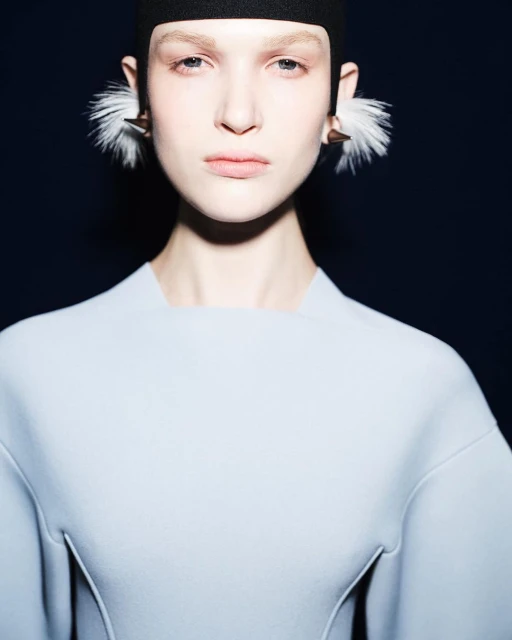 Close-up shot of a fair-skinned woman in a light blue dress with an intense gaze, featuring distinctive feathered earrings.