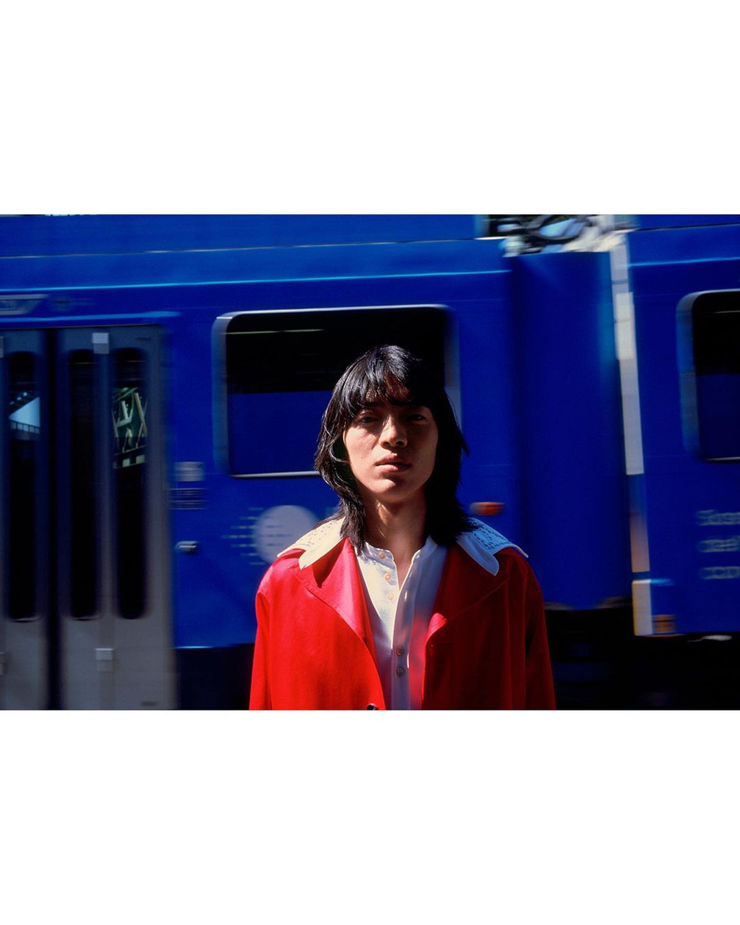 Adult stands in front of blurred blue train, wearing bright red jacket and white collared shirt. Medium outdoor shot with natural light.