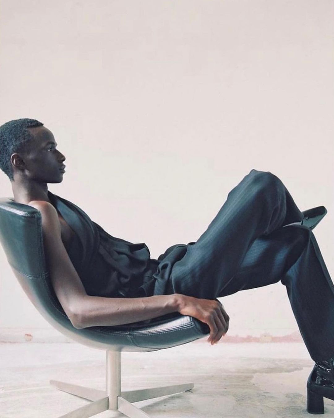 Adult male model reclines in a chair wearing a black top and pants in a studio with neutral wall. Fashion photography.