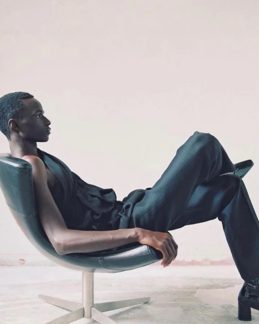 Adult male model reclines in a chair wearing a black top and pants in a studio with neutral wall. Fashion photography.
