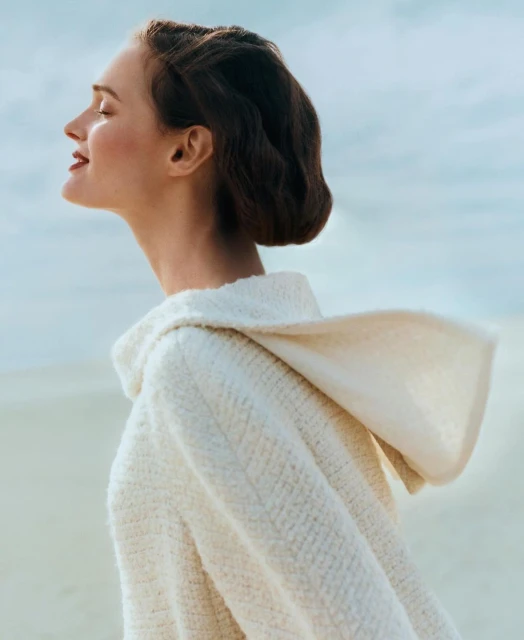 Woman in hooded knit sweater enjoying the light in an outdoor portrait against a diffused sky background in a fashion editorial style.