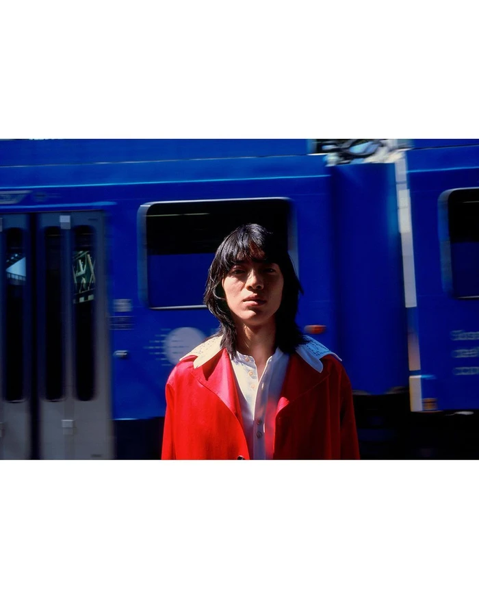 Model with long dark hair wearing a bright red jacket standing outdoors against a vibrant blue train background.