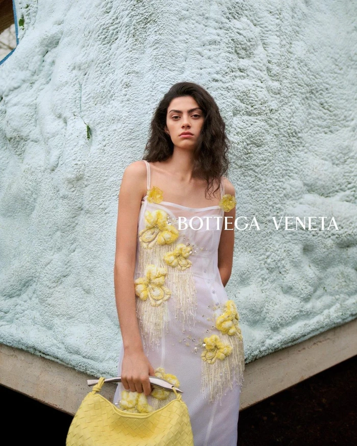 Model in white dress with yellow floral appliqués holding a yellow handbag against a textured foam wall