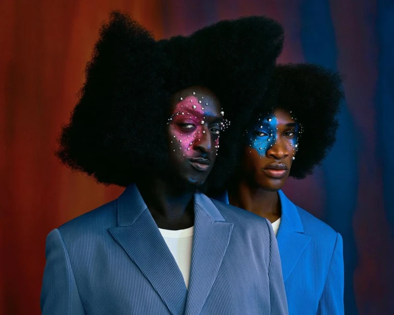 Two Black adults with colorful makeup and matching blue suits pose in front of a vibrant background. High-fashion editorial beauty shot.