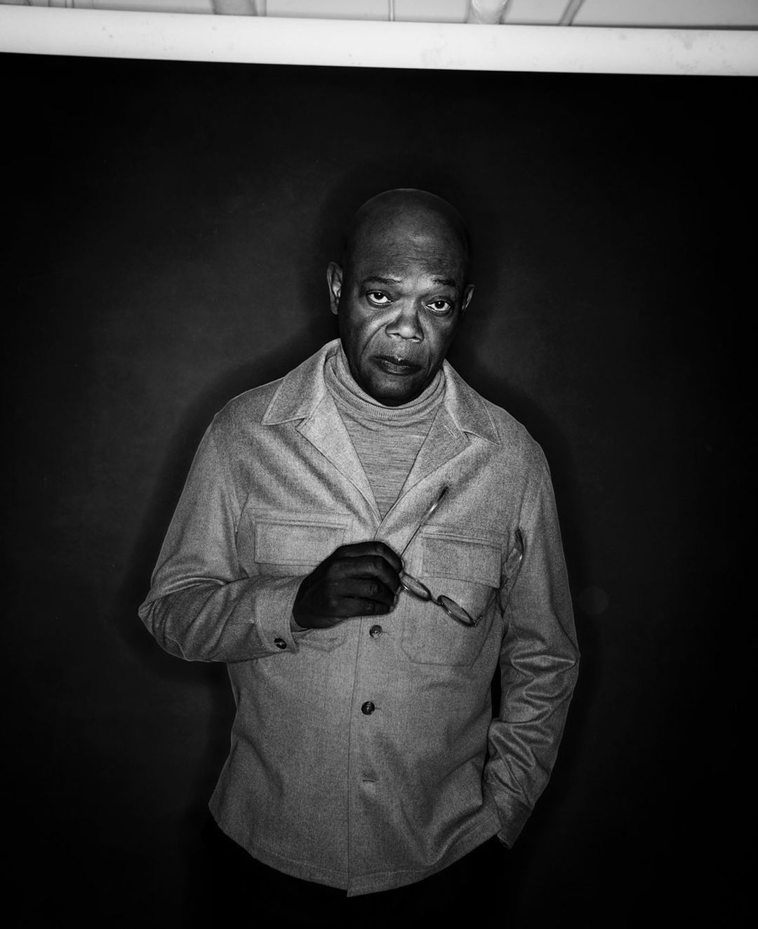 Black and white portrait of an adult man wearing a collared shirt and turtleneck, holding glasses against a dark background in studio.