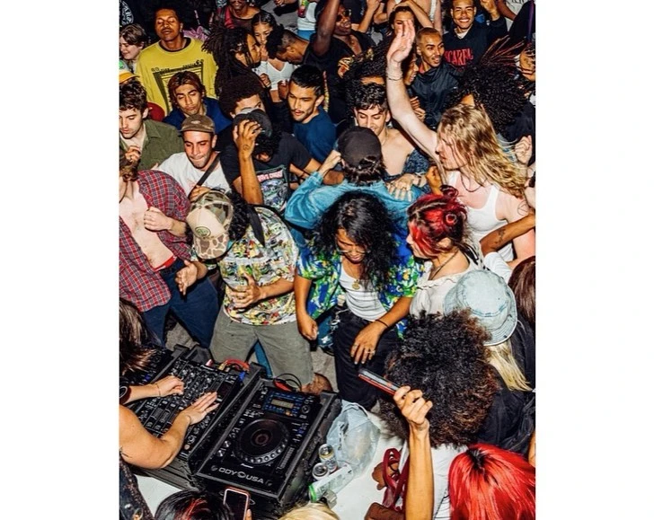 Energetic group of young adults dancing and enjoying a lively party, bathed in vibrant lighting with DJ equipment visible.