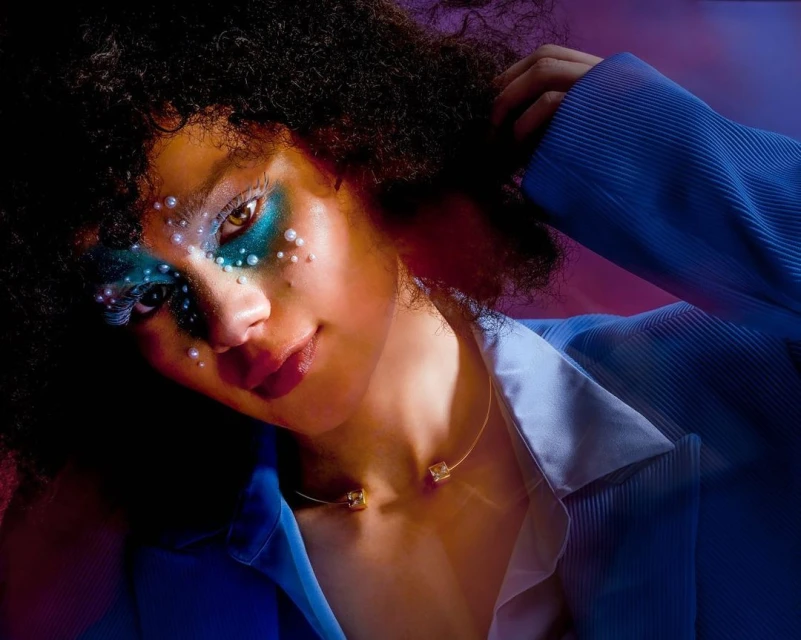 Close-up portrait of a young Black woman with Afro hair, wearing a blue jacket and pearls around her eyes, illuminated by purple light.