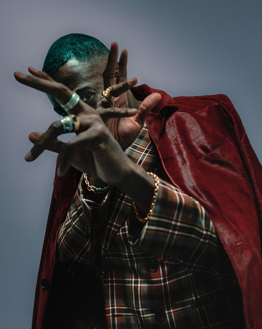 Portrait of a Black adult male wearing a plaid jacket and cape with rings, bracelets, and dyed teal hair, indoors with a gray background.