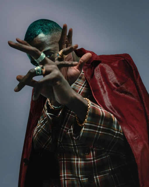 Portrait of a Black adult male wearing a plaid jacket and cape with rings, bracelets, and dyed teal hair, indoors with a gray background.