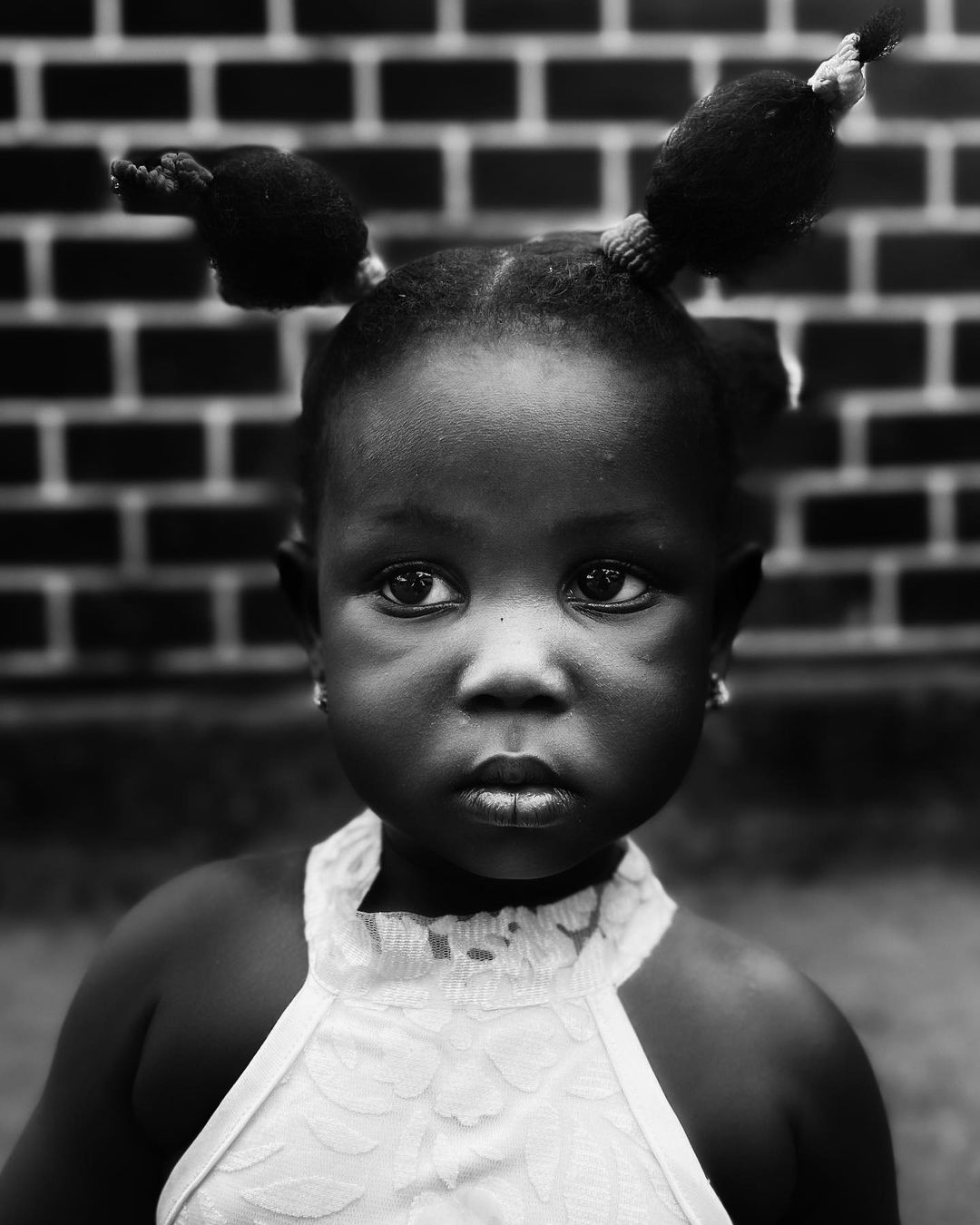 Portrait of a young Black girl with pigtails looking at the camera against a brick wall, captured in black and white with natural lighting.