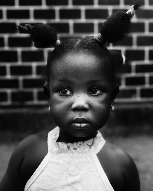 Portrait of a young Black girl with pigtails looking at the camera against a brick wall, captured in black and white with natural lighting.