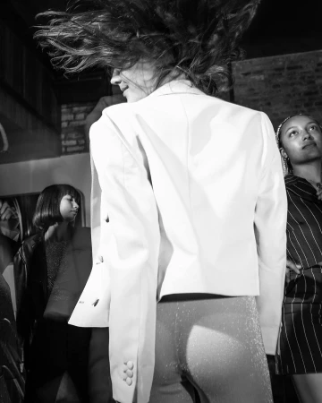 Monochrome photograph of a person wearing a light blazer with hair whipping in motion, flanked by two figures in an interior space.
