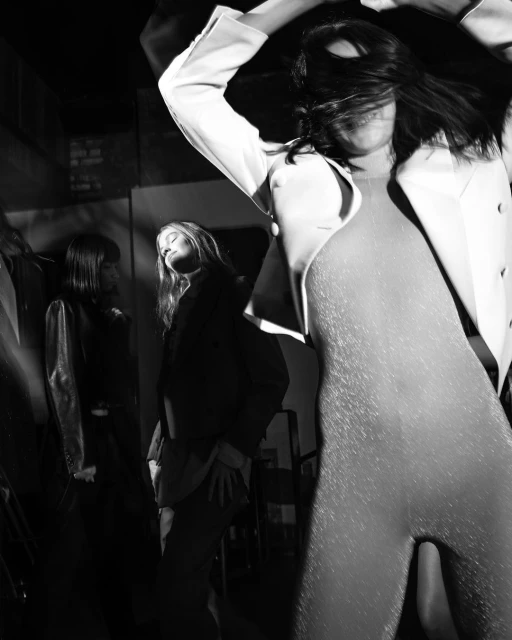 Three women pose in a black and white fashion photograph featuring a model in a shimmery catsuit and white blazer, expressing style and confidence.
