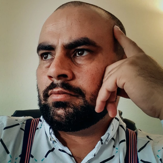Close-up portrait of a pensive adult male with a beard, wearing suspenders and a patterned shirt indoors with neutral lighting.