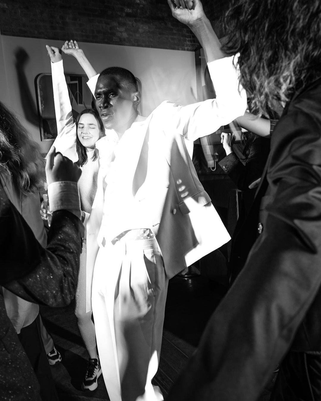 Monochrome image captures a group of people, including a man in a light suit, raising their hands in a lively indoor setting.