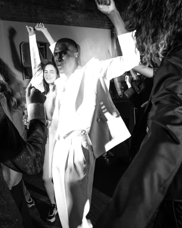 Monochrome image captures a group of people, including a man in a light suit, raising their hands in a lively indoor setting.