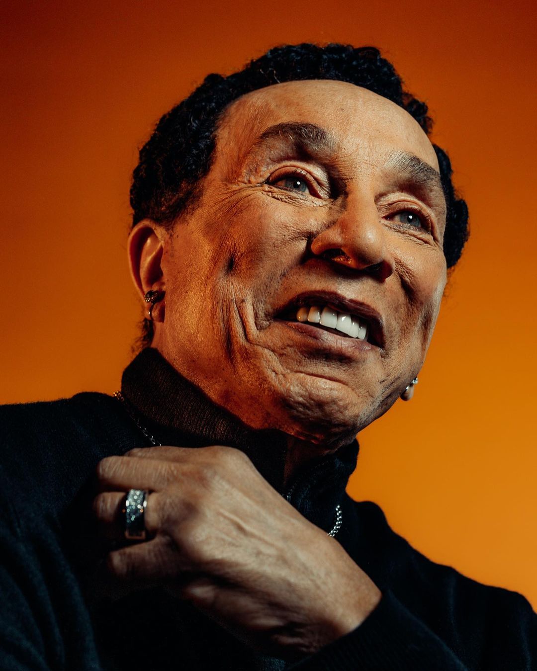 Close-up portrait of a mature Black man with a black turtleneck sweater against a warm orange background in a studio setting.