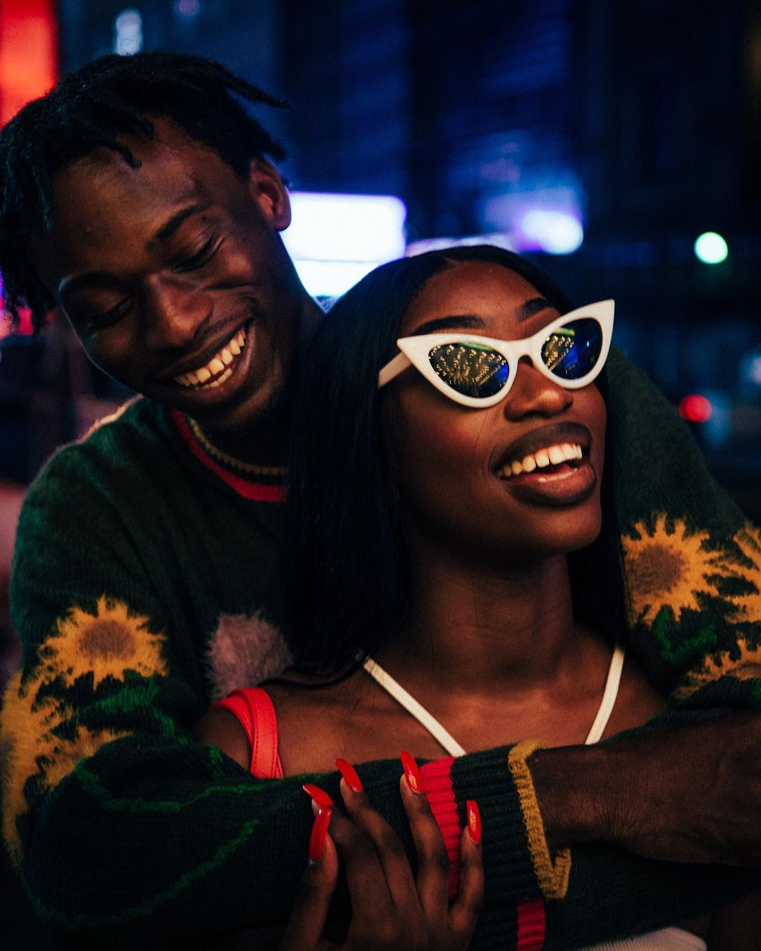 Smiling couple poses outdoors at night; a Black woman with white cat-eye sunglasses and red nails is embraced by a Black man.