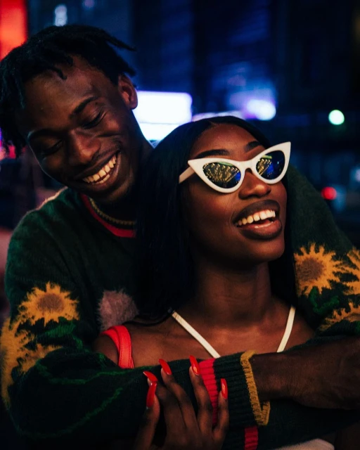 Smiling couple poses outdoors at night; a Black woman with white cat-eye sunglasses and red nails is embraced by a Black man.