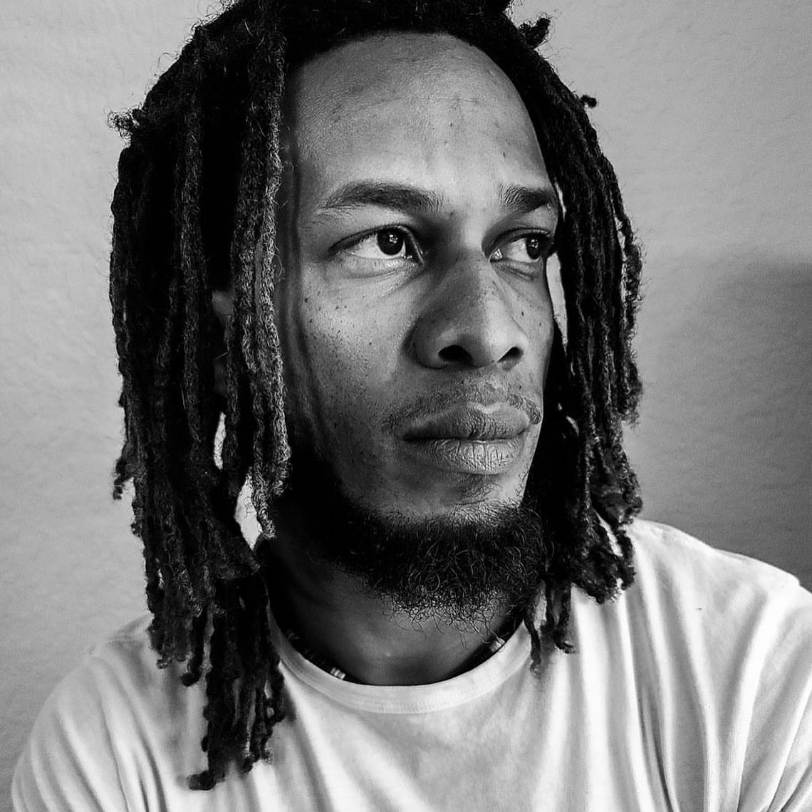 Portrait of a Black man with long dreadlocks and a beard wearing a light t-shirt, photographed by Malike Sidibe.