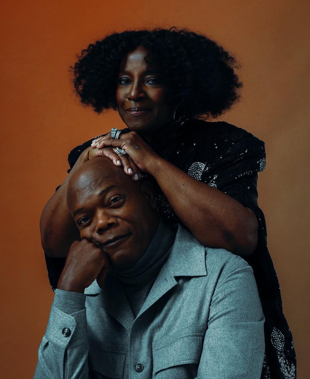 Portrait of Samuel L. Jackson with a Black woman in a black sequin dress, both smiling against a warm orange-toned backdrop.