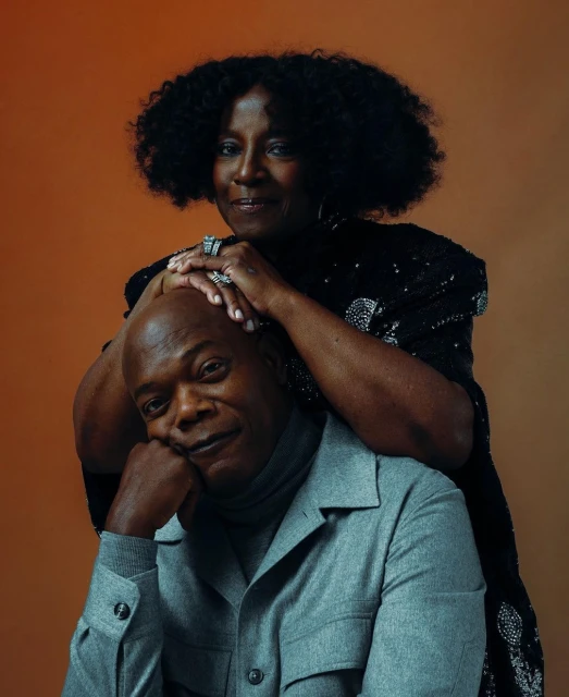 Portrait of Samuel L. Jackson with a Black woman in a black sequin dress, both smiling against a warm orange-toned backdrop.
