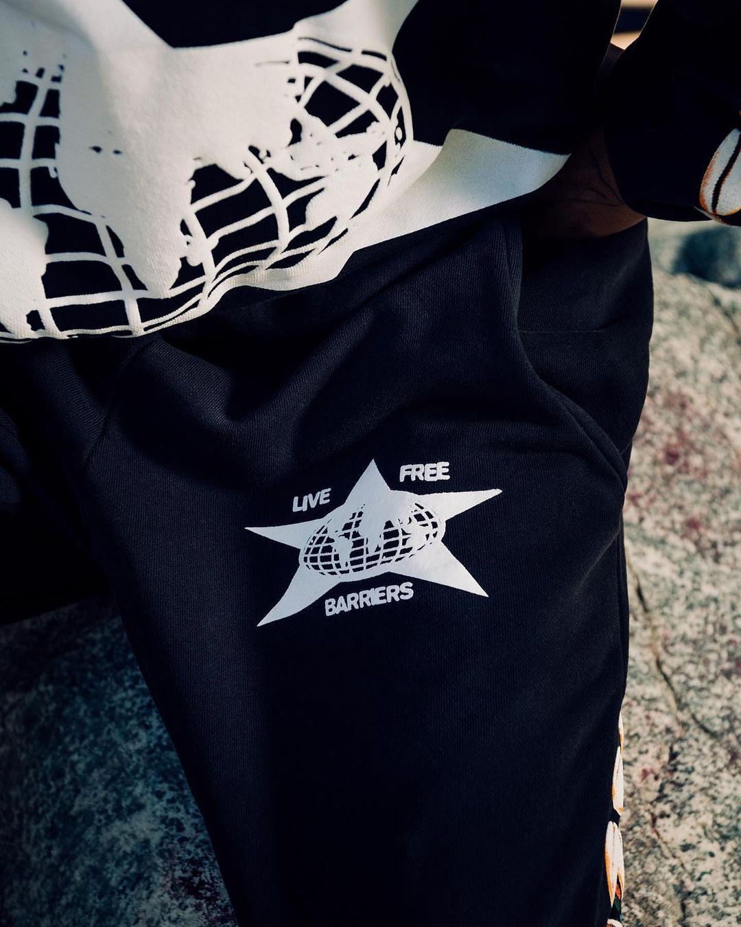 Close-up of person wearing black sweatpants with the white logo 'Live Free Barriers', along with a cream sweatshirt with a globe graphic.