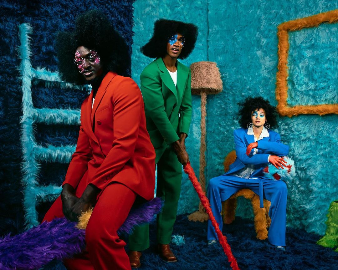 Three people in color-block suits pose in a surreal, textured room with a ladder, floor lamp, picture frame, and decorative furry seat.