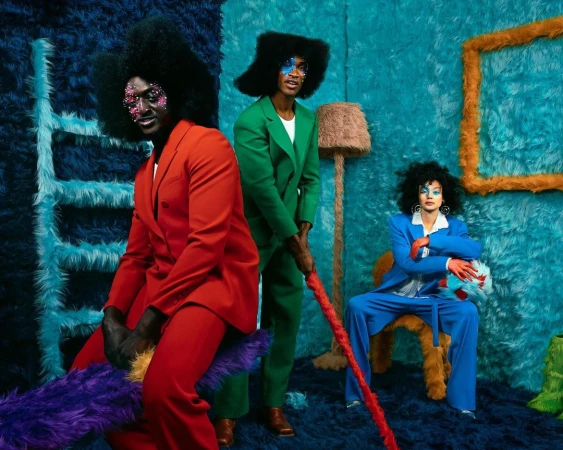 Three people in color-block suits pose in a surreal, textured room with a ladder, floor lamp, picture frame, and decorative furry seat.