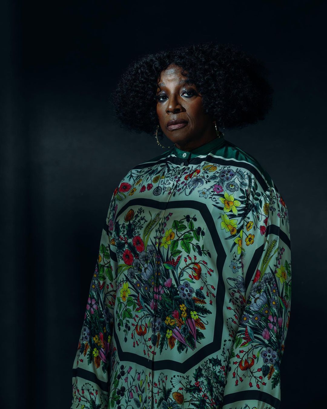 Portrait of an older Black woman wearing a patterned floral silk top against a dark moody background, photographed by Malike Sidibe.