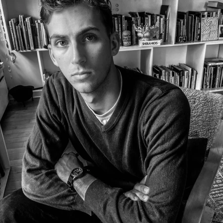 Portrait of a man in a dark v-neck sweater with arms crossed seated indoors by a bookshelf
