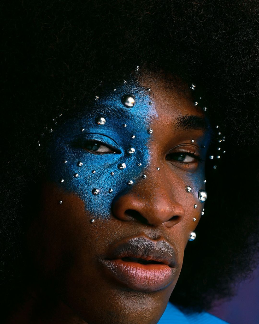Close-up of an adult with dark skin, striking blue face paint, and shiny silver embellishments, creating a bold, artistic beauty statement.