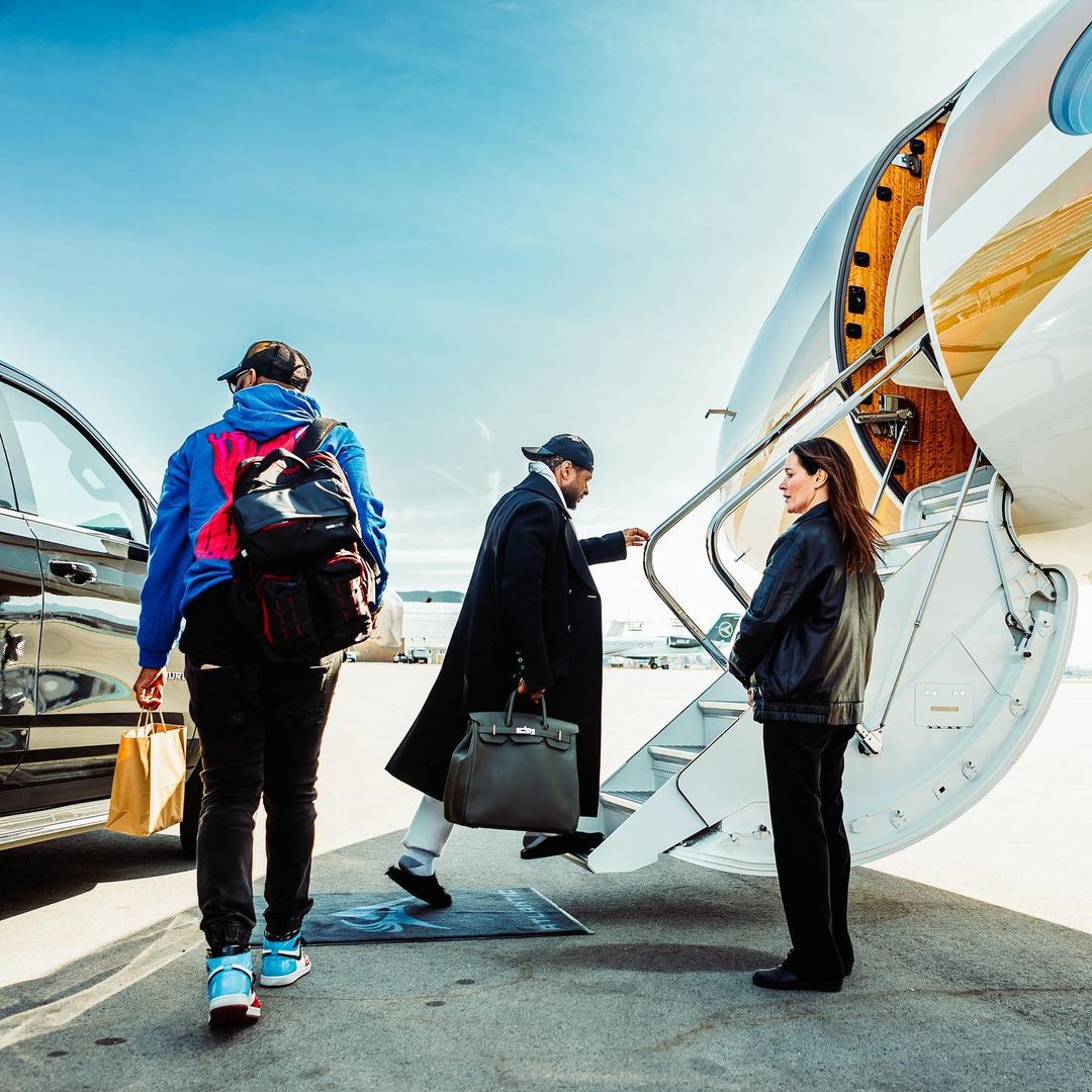 Two men board a private jet with a woman standing nearby on a sunny day. One man is carrying a bag and wearing a blue and pink jacket.