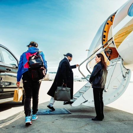 Two men board a private jet with a woman standing nearby on a sunny day. One man is carrying a bag and wearing a blue and pink jacket.