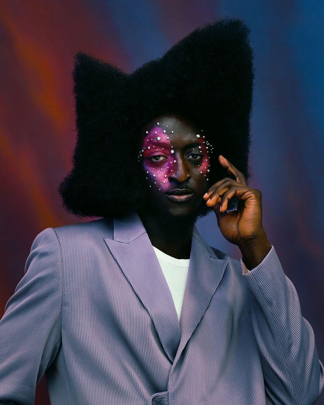 Portrait of a Black adult male with large afro, creative face makeup, wearing a striped lavender suit against a blurred studio backdrop.