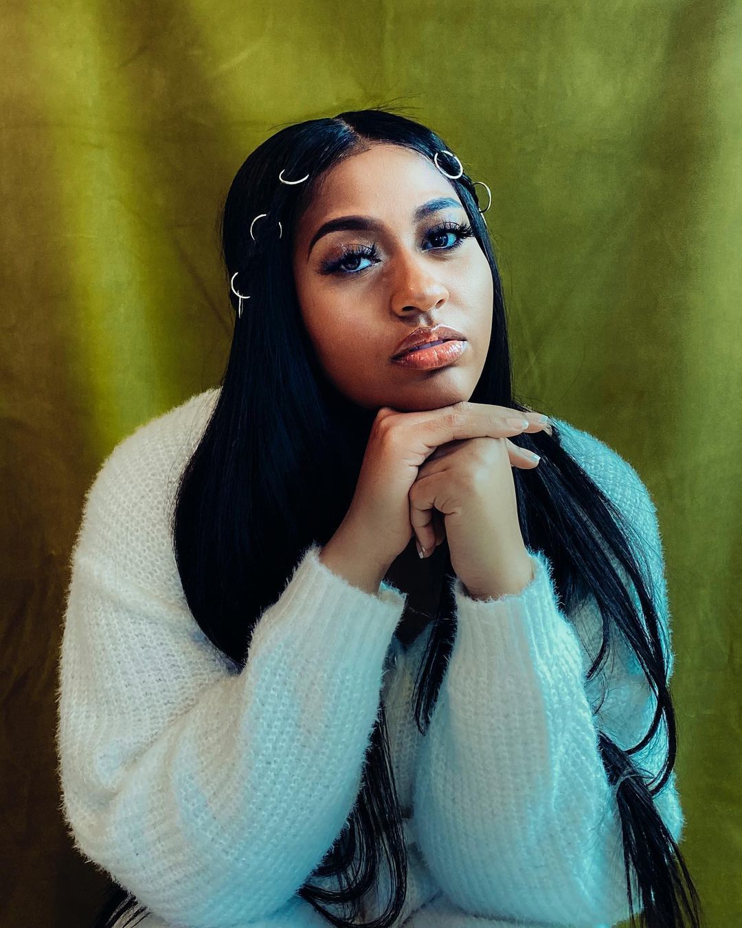 Portrait of a woman with long black hair wearing a white sweater with a green fabric background. She has hands near her face.
