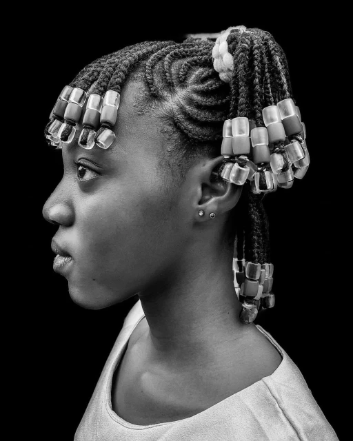 Profile portrait of a young Black woman with braided hair decorated with beads against a dark background, showcasing hairstyle.