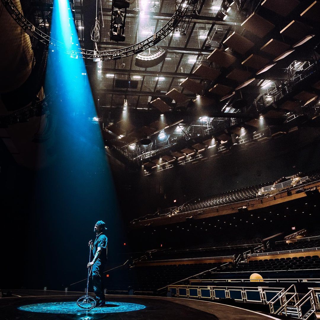 Man standing center stage under a bright blue spotlight in a large, empty theater; stands next to a unicycle. The stage is dark.