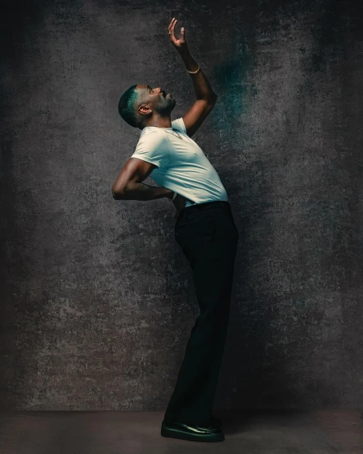 A Black male in a white t-shirt and black pants poses against a textured grey backdrop with his arm raised and body arched backward.