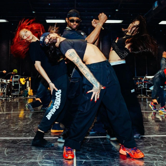Group of four people dancing indoors, with the central figure leaning back showing a tattooed stomach. There are guitars and stage equipment in the background.
