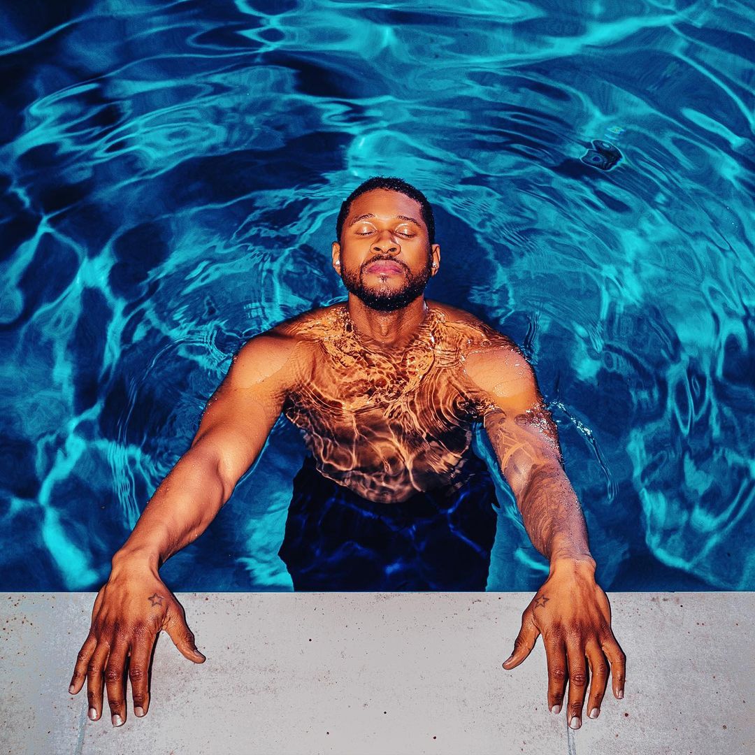A man with a beard and short hair leans against the edge of a blue-tiled pool with sunlight rippling across his shoulders and chest.