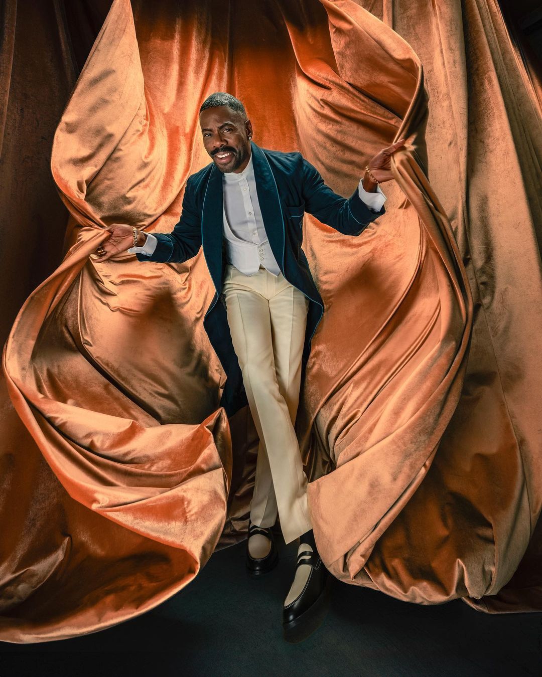 Adult male in an unbuttoned, teal tailcoat and cream-colored pants stands smiling amid copper-colored velvet drapes in a studio shot.