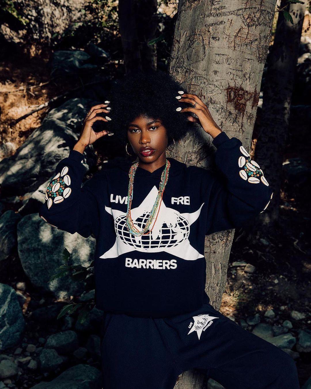 A woman with afro stands next to a tree trunk in a black matching set. Her hands are on her head, and she wears a beaded necklace.