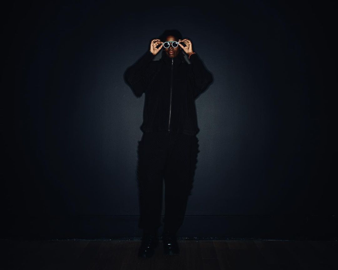 Person in a black outfit stands against a dark gray background, holding round sunglasses up to their face.