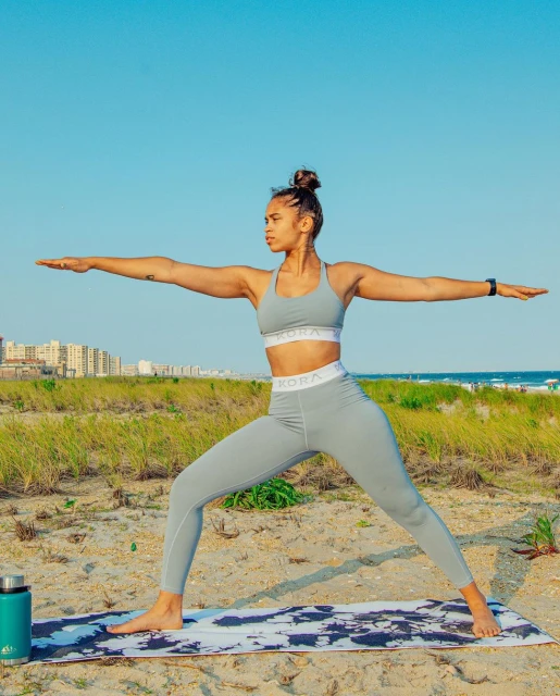 Athletic woman in matching activewear performs Warrior II pose on a yoga mat on the beach with buildings and blue sky in the background.