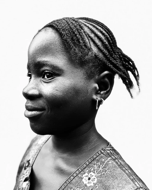 Portrait of a young African-descent woman with braided hair and a denim top, smiling slightly against a bright, neutral background.